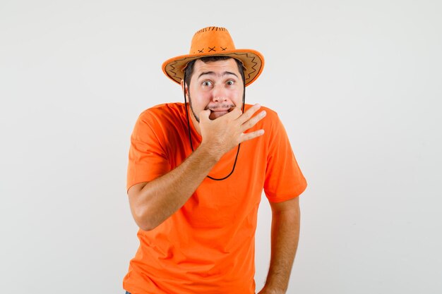 Jeune homme sifflant en t-shirt orange, chapeau et semblant anxieux. vue de face.