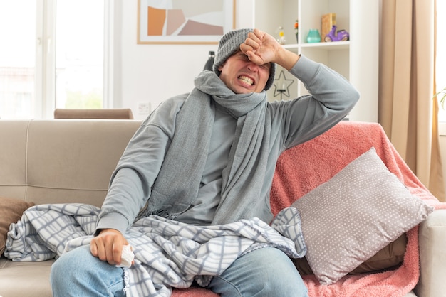 Jeune homme slave malade avec une écharpe autour du cou portant un chapeau d'hiver mettant la main sur le front et tenant un tissu assis sur un canapé dans le salon