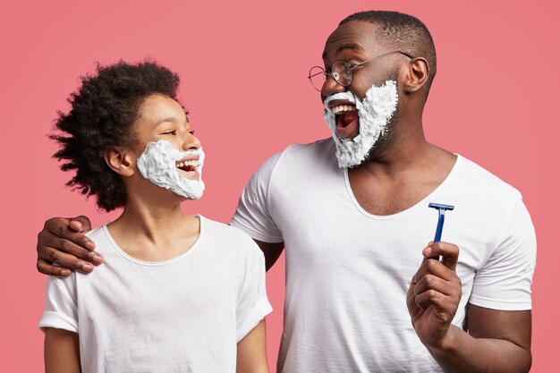 Jeune homme et son fils ayant de la mousse à raser sur le visage et tenant des rasoirs