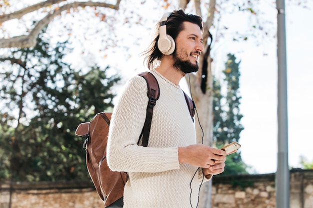 Photo gratuite jeune homme avec son sac à dos, écoute de musique sur téléphone portable à travers des écouteurs à l'extérieur