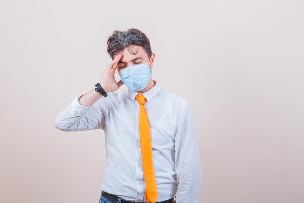 Jeune homme souffrant de maux de tête en chemise, cravate, jeans, masque et ayant l'air épuisé