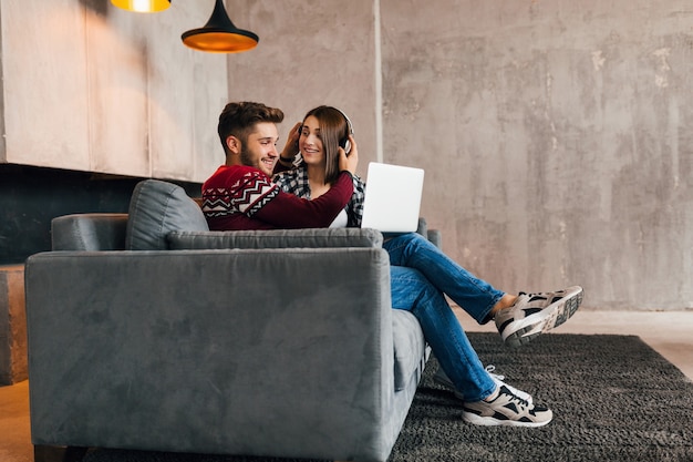 Jeune homme souriant assez heureux et femme assise à la maison en hiver, à la recherche d'un ordinateur portable, à l'écoute d'un casque, les étudiants qui étudient en ligne, couple sur le temps libre