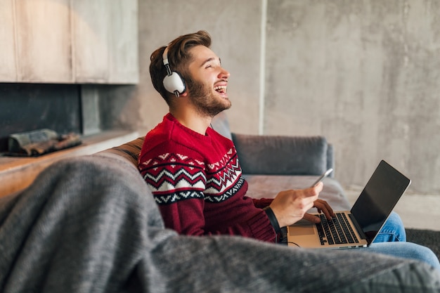 Jeune Homme Souriant Attrayant Sur Le Canapé à La Maison En Hiver Chantant De La Musique Sur Des écouteurs, Portant Un Pull En Tricot Rouge, Travaillant Sur Ordinateur Portable, Pigiste, émotionnel, Riant, Heureux