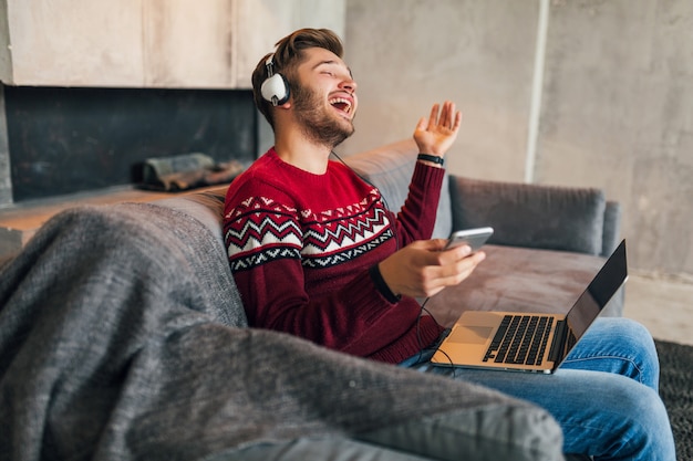 Jeune homme souriant attrayant sur le canapé à la maison en hiver chantant de la musique sur des écouteurs, portant un pull en tricot rouge, travaillant sur ordinateur portable, pigiste, émotionnel, rire, heureux