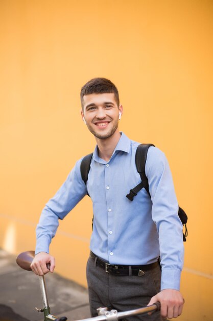 Jeune homme souriant en chemise bleue et sac à dos debout avec vélo regardant joyeusement de côté sur fond orange en plein air