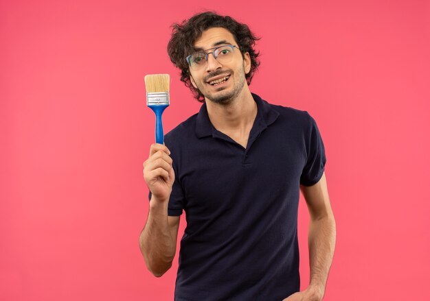 Jeune homme souriant en chemise noire avec des lunettes optiques détient un pinceau isolé sur un mur rose