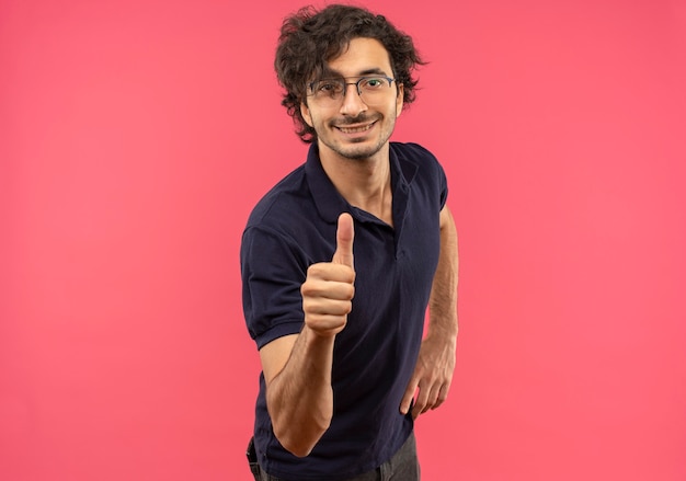 Jeune homme souriant en chemise noire avec des lunettes optiques pouces vers le haut et semble isolé sur un mur rose