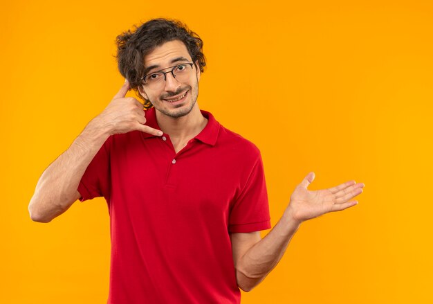 Jeune homme souriant en chemise rouge avec des gestes de lunettes optiques m'appellent signe de la main et points à côté isolé sur mur orange
