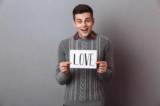 Jeune homme souriant, debout, isolé, tenant du papier avec le texte de l'amour.