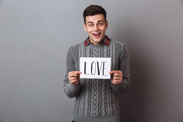 Jeune homme souriant, debout, isolé, tenant du papier avec le texte de l'amour.