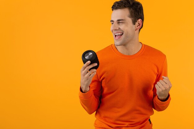 Jeune homme souriant élégant en pull orange tenant un haut-parleur sans fil heureux d'écouter de la musique en s'amusant