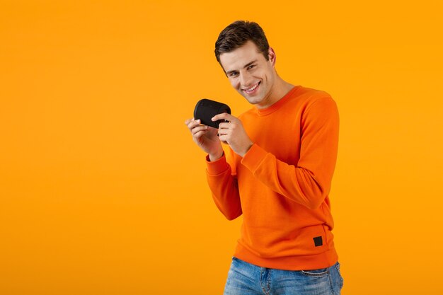 Jeune homme souriant élégant en pull orange tenant un haut-parleur sans fil heureux d'écouter de la musique en s'amusant