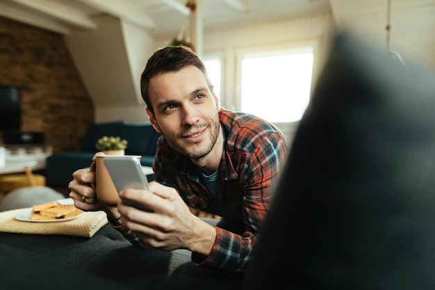 Jeune homme souriant envoyant des SMS sur son téléphone portable tout en se relaxant dans le salon et en buvant du café