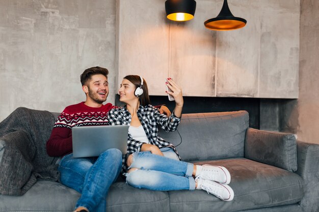 Jeune homme souriant et femme assise à la maison en hiver avec un ordinateur portable, écoutant des écouteurs, couple sur le temps libre ensemble, faisant selfie photo sur appareil photo smartphone, heureux, positif, rencontres, rire