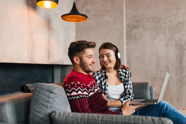 Jeune homme souriant et femme assise à la maison en hiver, travaillant sur ordinateur portable, écoutant des écouteurs, couple sur les loisirs, passer du temps ensemble, pigiste, heureux, rencontres