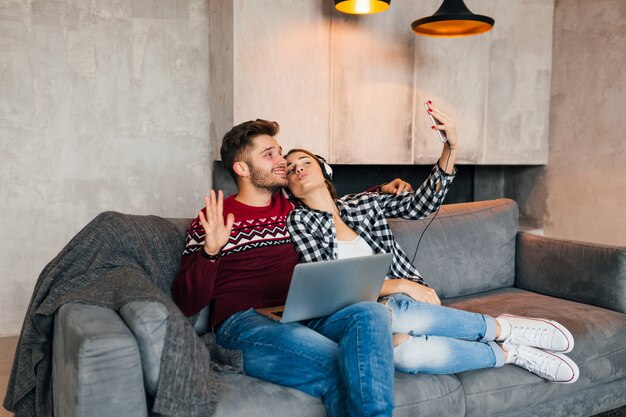 Jeune homme souriant et femme assise à la maison en hiver, travaillant sur ordinateur portable, tenant un smartphone, écoutant des écouteurs, couple sur les loisirs, passer du temps en ligne, pigiste, rencontres
