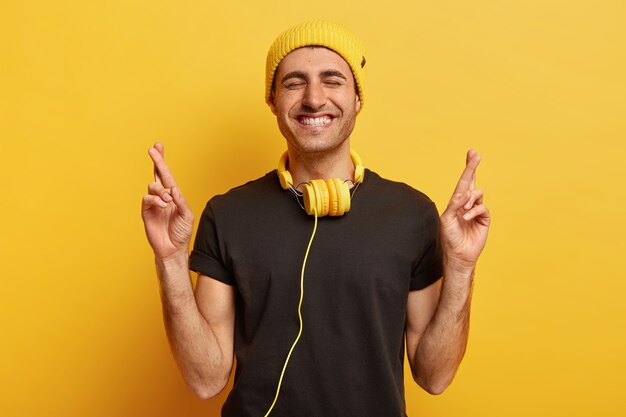 Un jeune homme souriant positif prie le rêve devenu réalité, croise les doigts et sourit largement, montre des dents blanches