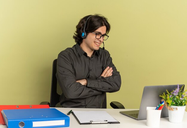 Jeune homme souriant de travailleur de bureau sur le casque dans des lunettes optiques est assis au bureau avec des outils de bureau à l'aide et en regardant un ordinateur portable avec les bras croisés isolé sur fond vert avec espace de copie