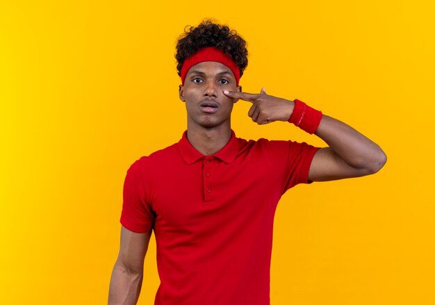 Jeune homme sportif afro-américain portant bandeau et bracelet mettant le doigt sur l'oeil isolé sur mur jaune