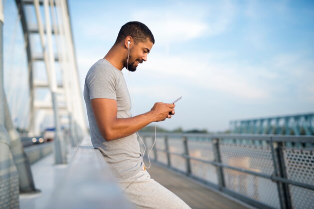 Jeune homme sportif attrayant à l'aide de téléphone et souriant sur le pont