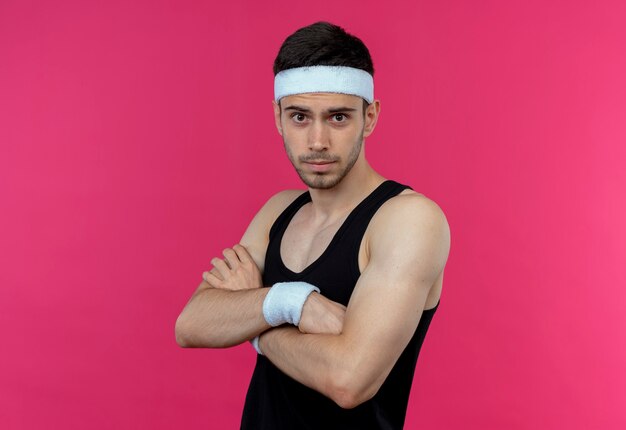 Jeune homme sportif en bandeau avec une expression sérieuse confiante avec les bras croisés sur la poitrine debout sur le mur rose