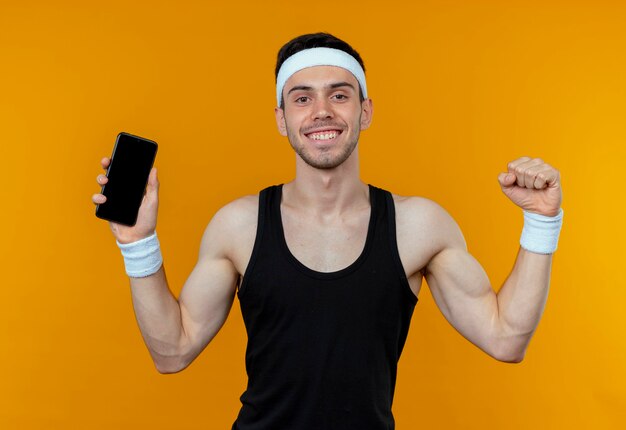 Jeune homme sportif en bandeau montrant le poing serrant le smartphone heureux et excité debout sur le mur orange