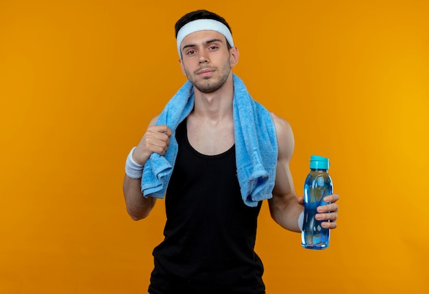 Jeune homme sportif en bandeau avec une serviette autour du cou tenant une bouteille d'eau regardant la caméra avec une expression confiante debout sur fond orange
