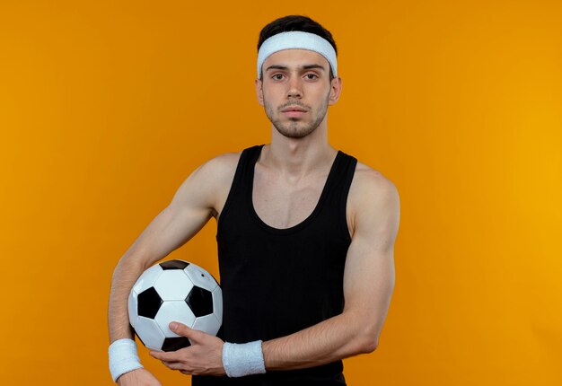 Jeune homme sportif en bandeau tenant un ballon de football avec une expression sérieuse confiante debout sur un mur orange