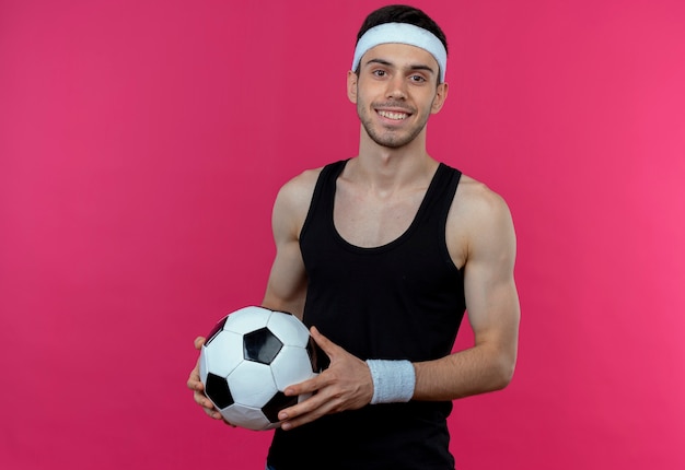 Jeune homme sportif en bandeau tenant un ballon de football souriant joyeusement debout sur un mur rose