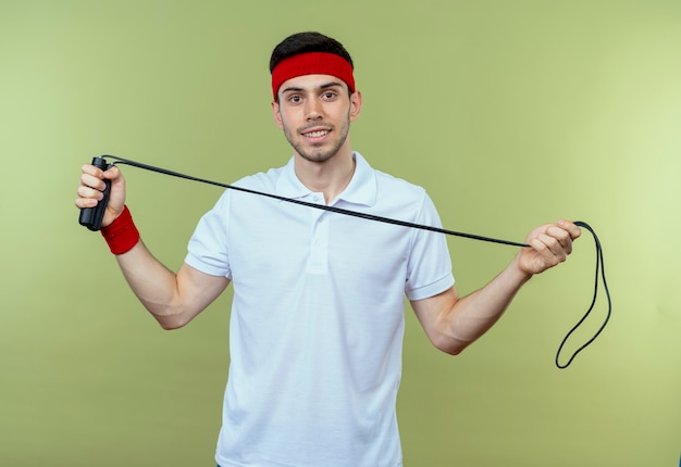 Jeune homme sportif en bandeau tenant la corde à sauter souriant sur vert