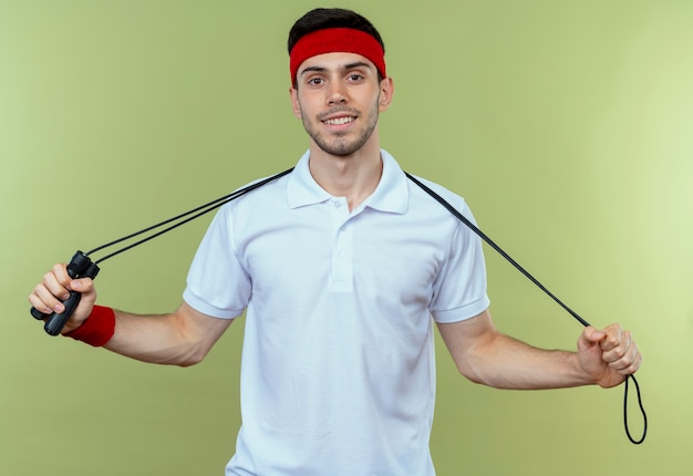 Photo gratuite jeune homme sportif en bandeau tenant la corde à sauter avec le sourire sur le visage sur vert