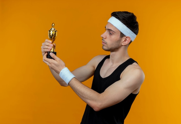 Photo gratuite jeune homme sportif en bandeau tenant son trophée en le regardant avec une expression sérieuse confiante debout sur un mur orange