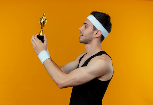 Jeune homme sportif en bandeau tenant son trophée en regardant ithappy and positive smiling standing over orange wall