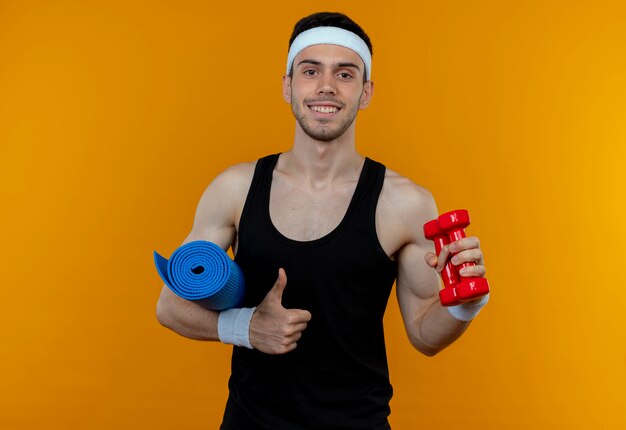 Jeune homme sportif en bandeau tenant tapis de yoga et dumbbel montrant les pouces vers le haut souriant joyeusement debout sur le mur orange