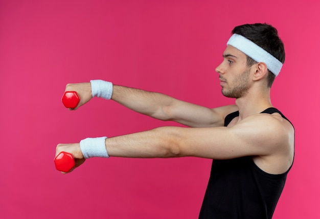 Jeune homme sportif en bandeau travaillant avec des haltères tendus et confiant debout sur fond rose