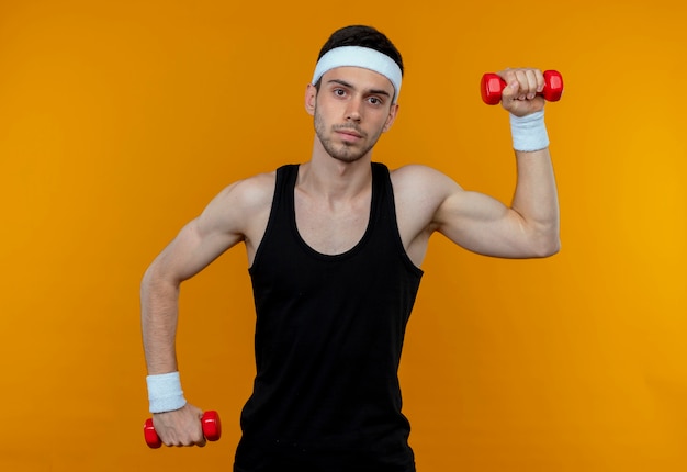 Jeune homme sportif en bandeau travaillant avec des haltères avec un visage sérieux sur orange