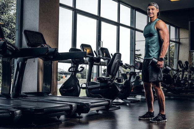 Jeune homme sportif fait des exercices avec des haltères dans un club de gym vide.