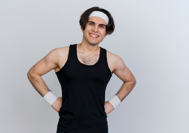 Jeune homme sportif portant des vêtements de sport et un bandeau à l'avant heureux et positif souriant joyeusement debout sur un mur blanc