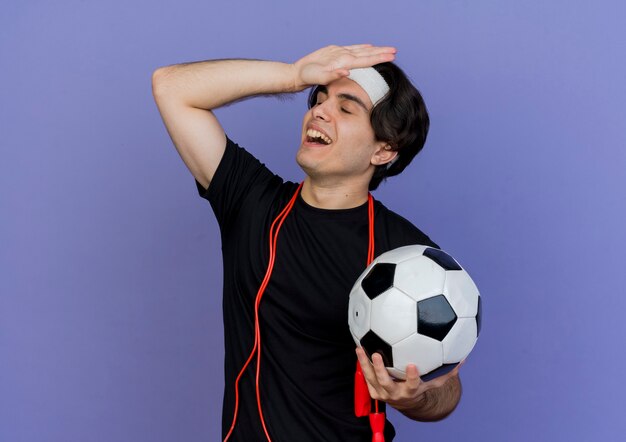 Jeune homme sportif portant des vêtements de sport et un bandeau avec une corde à sauter autour du cou tenant un ballon de football à la confusion avec la main sur sa tête pour erreur
