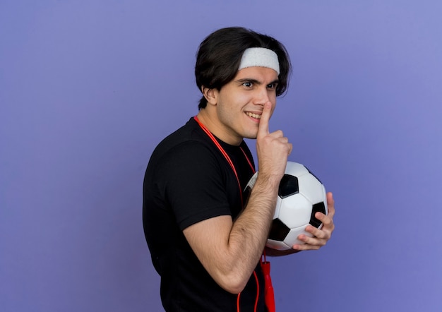 Jeune homme sportif portant des vêtements de sport et un bandeau avec une corde à sauter autour du cou tenant un ballon de football faisant un geste de silence avec le doigt sur les lèvres