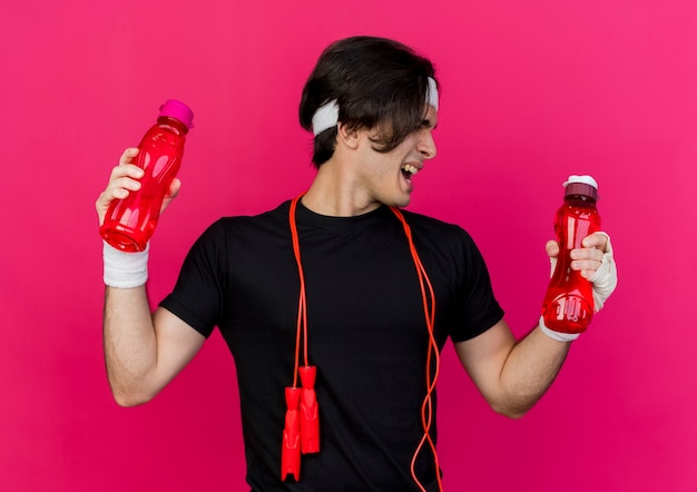 Photo gratuite jeune homme sportif portant des vêtements de sport et un bandeau avec une corde à sauter autour du cou tenant deux bouteilles d'eau en regardant une bouteille avec une expression agacée