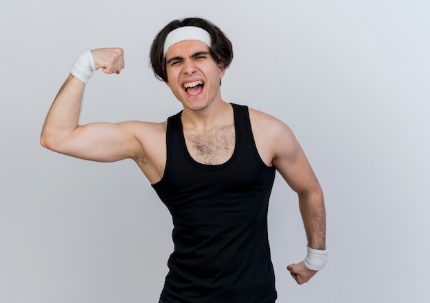 Jeune homme sportif portant des vêtements de sport et un bandeau levant le poing montrant la force et les biceps à la tension debout sur un mur blanc