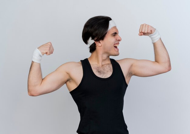 Jeune homme sportif portant des vêtements de sport et un bandeau levant les poings montrant la force et les biceps à la tension debout sur un mur blanc