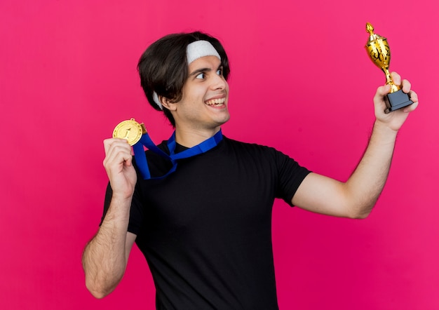 Photo gratuite jeune homme sportif portant des vêtements de sport et un bandeau avec médaille d'or autour du cou montrant son trophée heureux et excité en le regardant