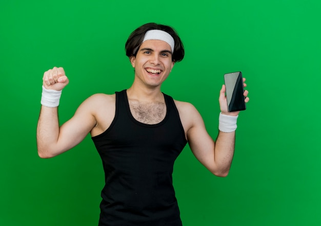 Jeune homme sportif portant des vêtements de sport et un bandeau montrant le poing serrant le smartphone heureux et excité