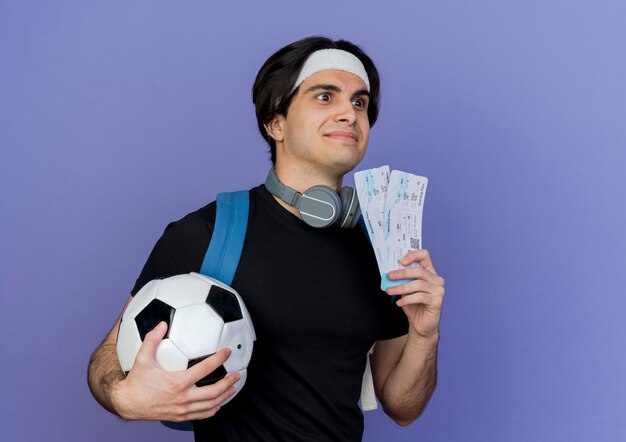 Jeune homme sportif portant des vêtements de sport et un bandeau avec sac à dos tenant un ballon de football et des billets d'avion à côté souriant confus