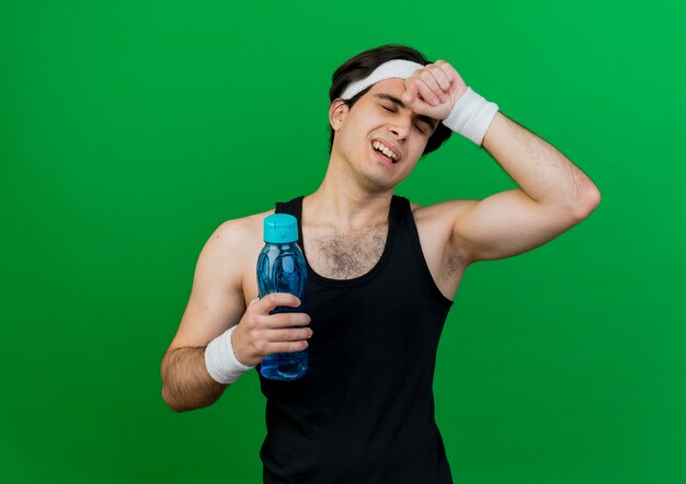 Jeune homme sportif portant des vêtements de sport et bandeau tenant une bouteille d'eau à la fatigue