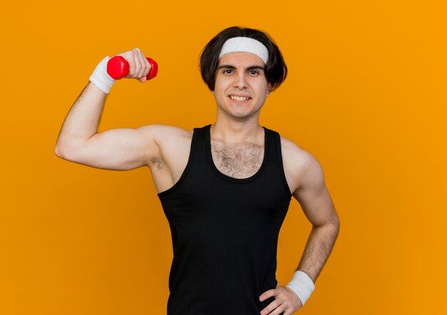 Jeune homme sportif portant des vêtements de sport et un bandeau tenant un haltère en main levée faisant des exercices souriant debout confiant