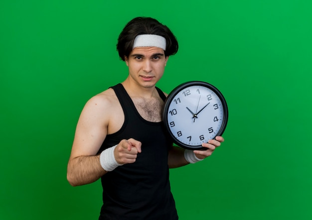 Jeune homme sportif portant des vêtements de sport et un bandeau tenant une horloge murale pointant avec l'index à la caméra avec un visage sérieux