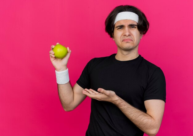 Jeune homme sportif portant des vêtements de sport et un bandeau tenant une pomme verte le présentant avec une expression triste sur le visage debout